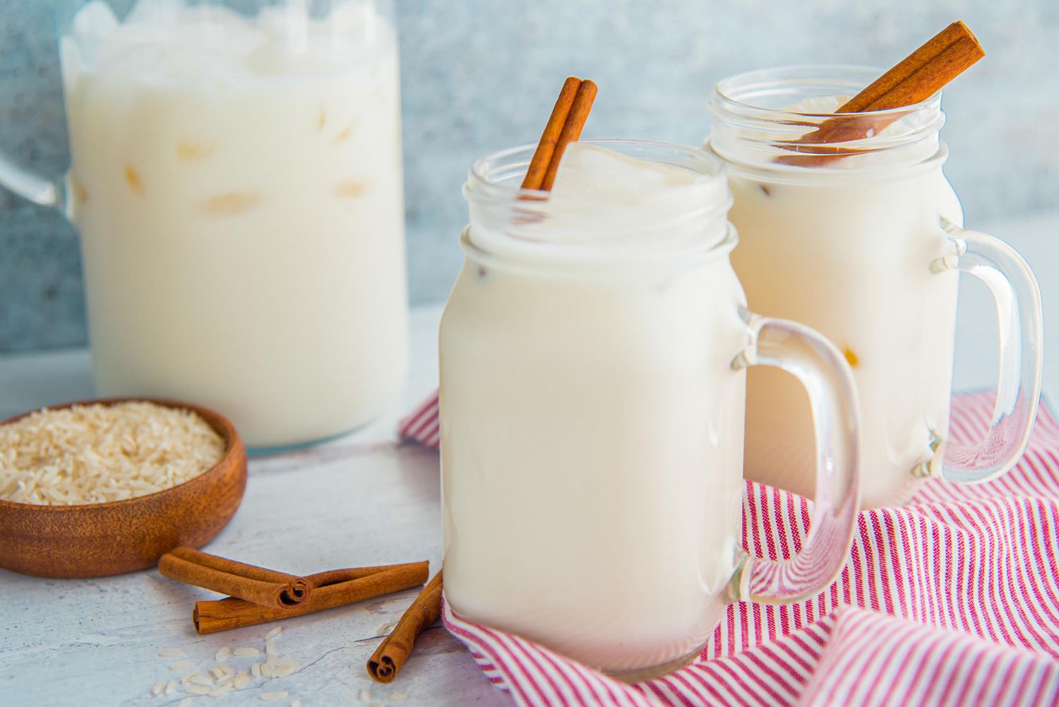 A large mason jar of easy homemade horchata with a cinnamon stick inside is in front of a similar glass and a pitcher of homemade horchata. A small bowl of rice and cinnamon sticks are to the left.