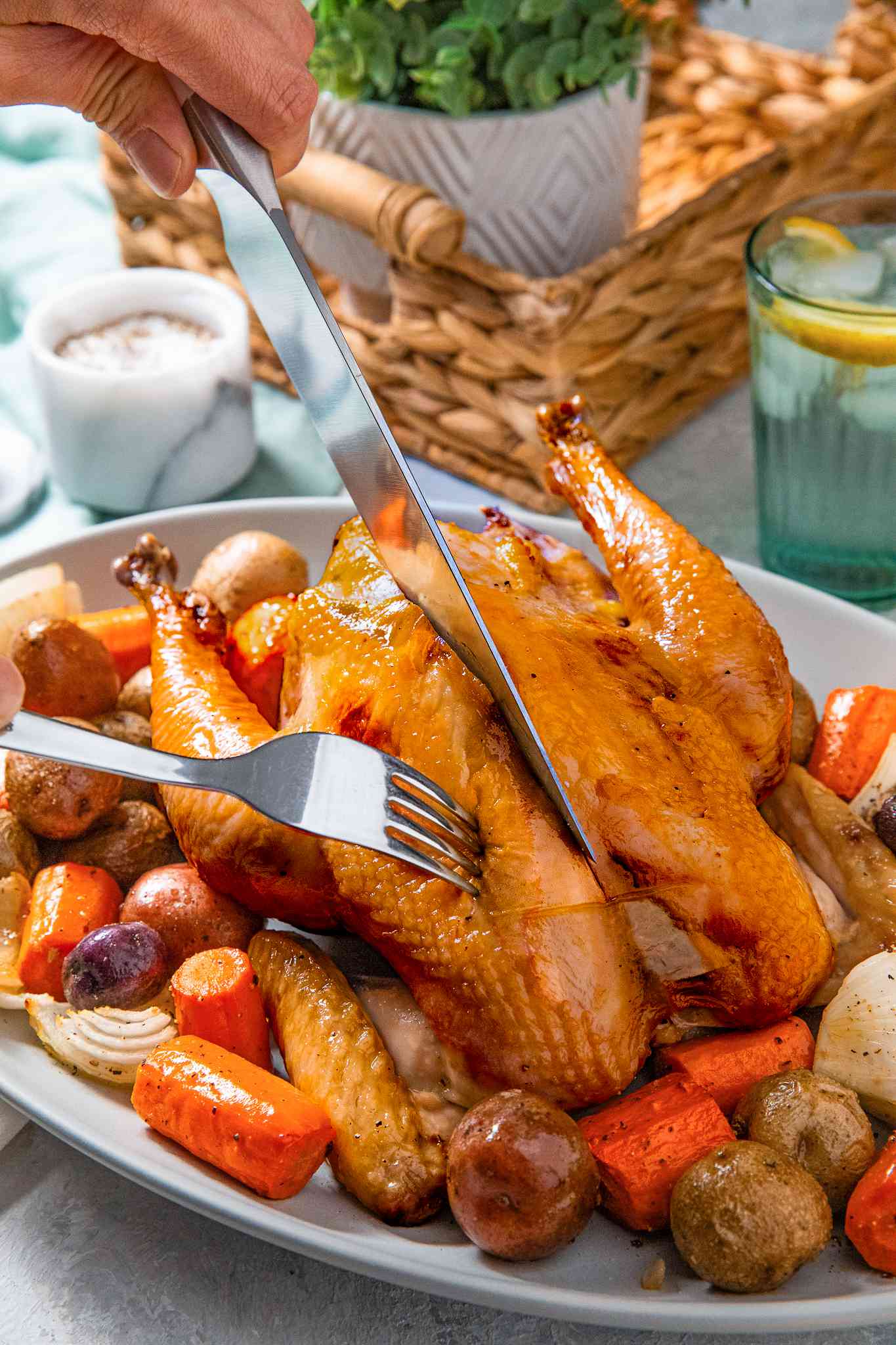 Carving knife cutting into simple roast chicken that's served on a bed of roasted vegetables at a table setting with plates, a basket with a potted plant, a glass, and utensils 